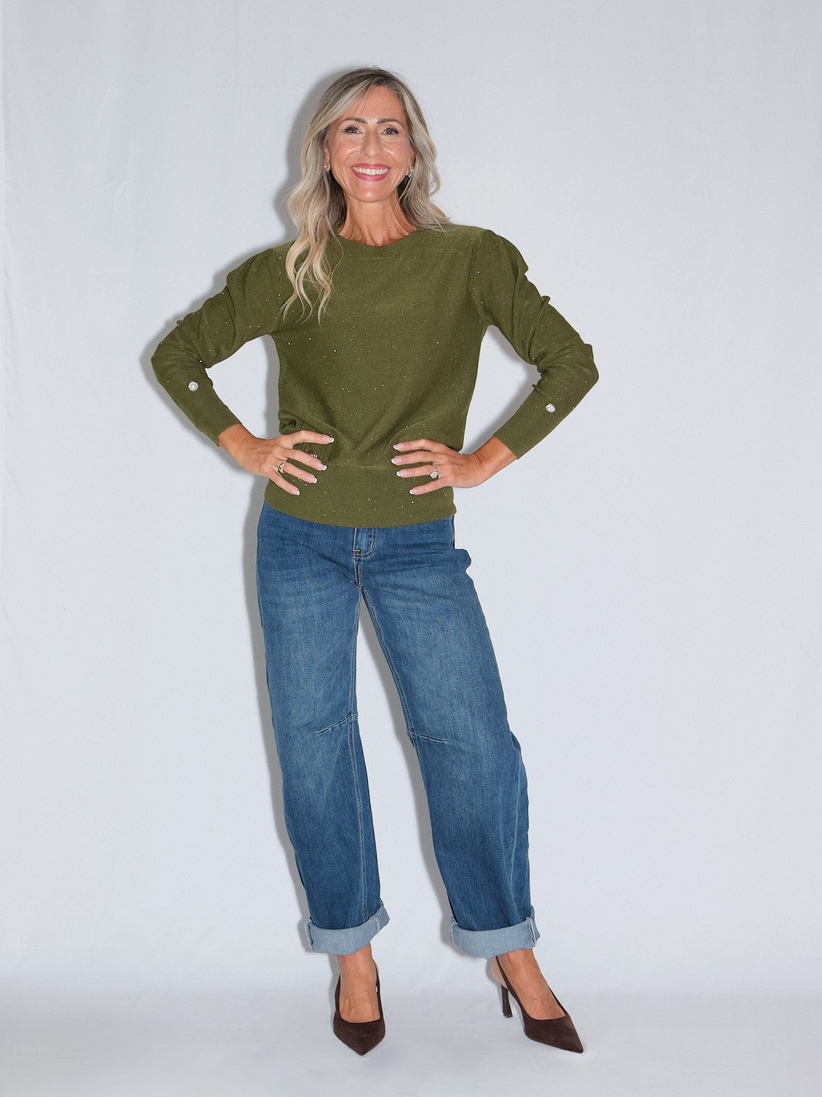 Un modello che indossa un girocollo manica lunga lurex verde con dettagli di bottoncini, abbinato a jeans e scarpe eleganti.